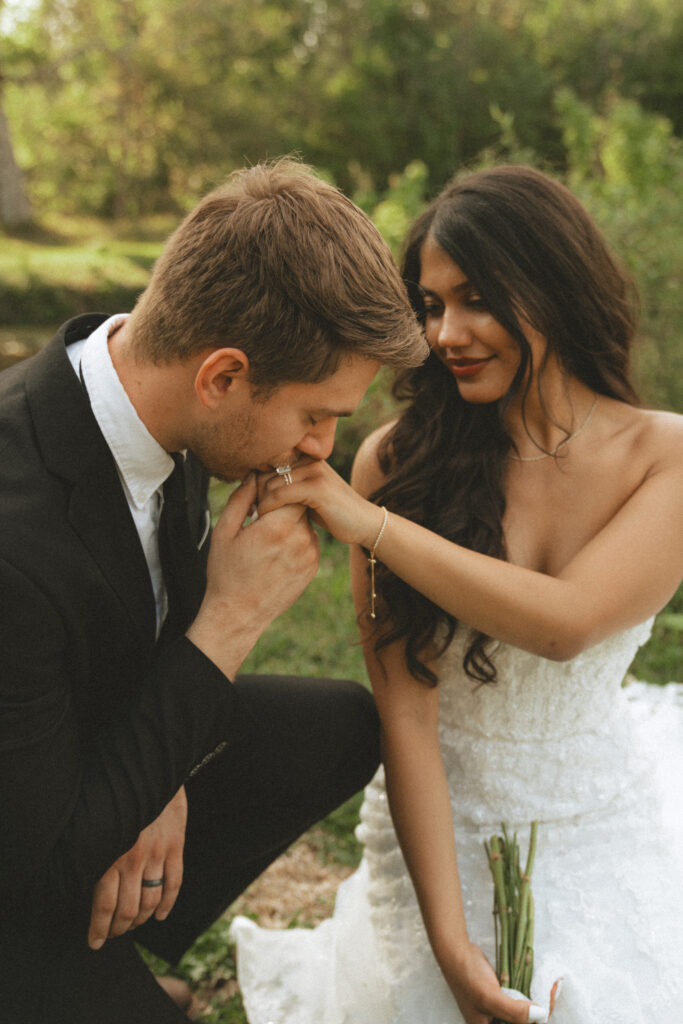 groom kissing his brides hand as she sits on the grass and smiles at him