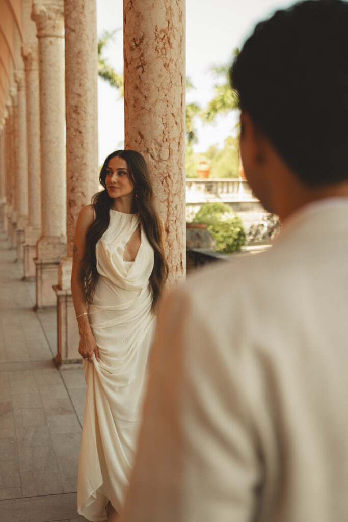 groom looks at his bride as she leans on a column at the Ringling museum for their engagement photos