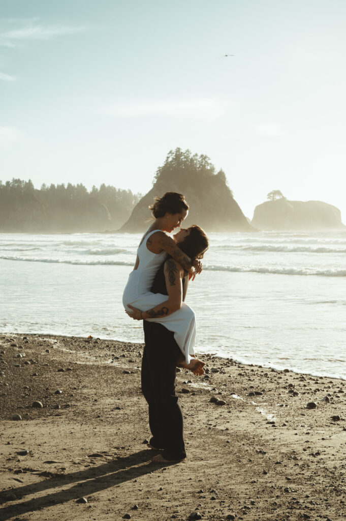 one bride holding the other in her arms on the beach