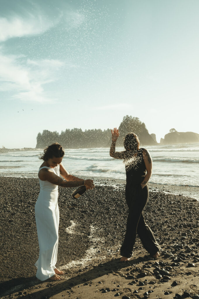 one bride popping a bottle of champagne as it sprays at her wife with rialto beach in the background