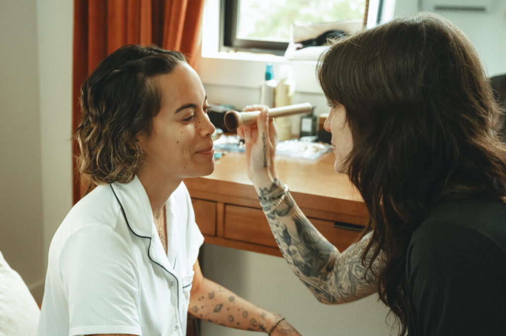 One bride doing the other brides makeup in their room on the morning of their wedding