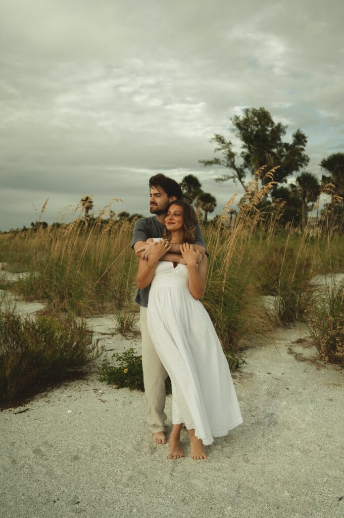 man hugging a woman from behind as they look out at the ocean on Pass-a-grille beach for their engagement photos