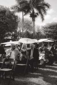 a candid photo of guests sitting at a wedding ceremony with parasols, waiting for the bride